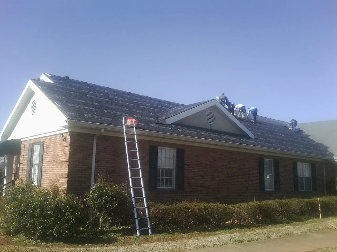 Crew tearing shingles off a brick ranch roof with a ladder set — Emergency Roof Repair work by our crew in The Colony